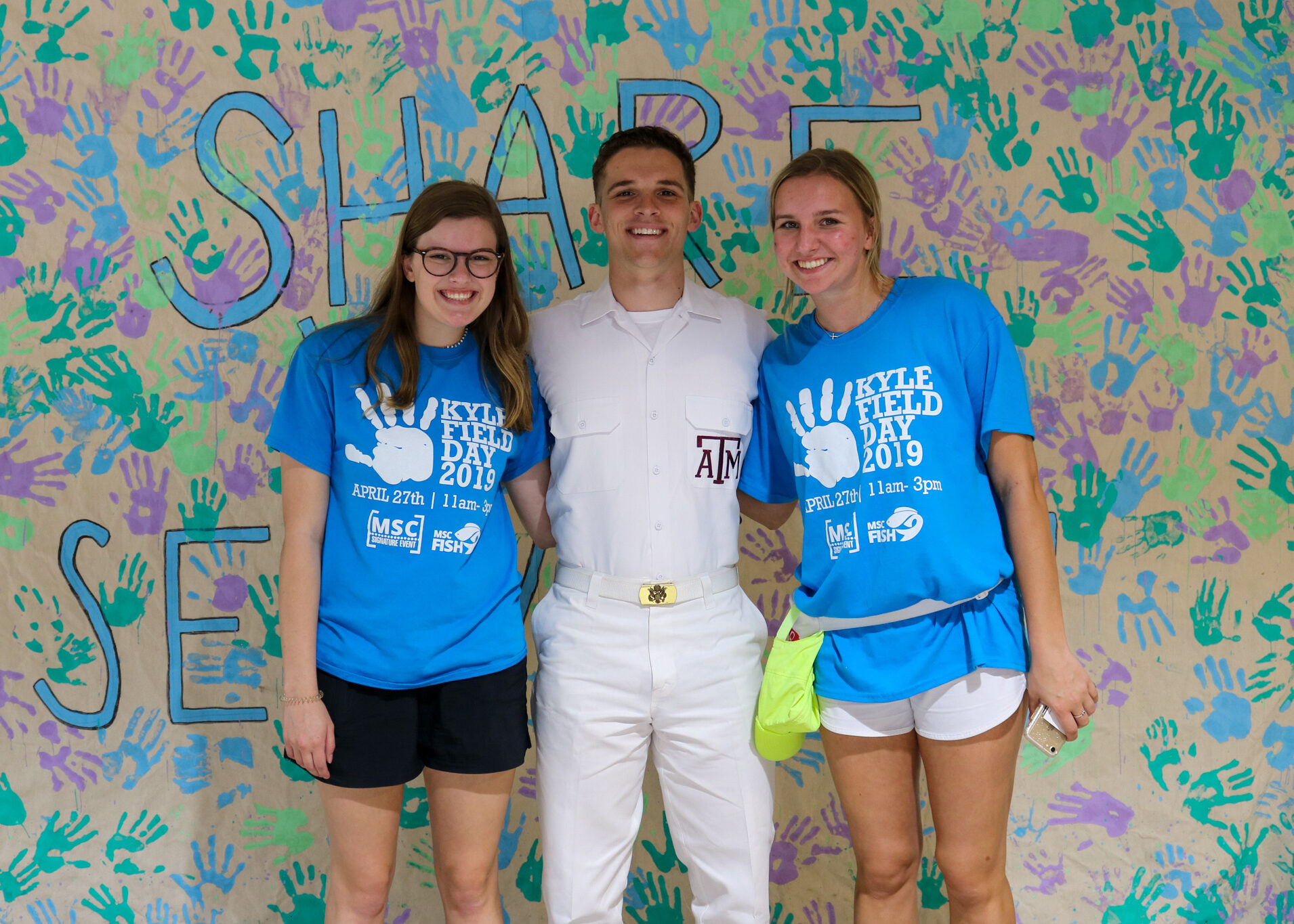 Two Kyle Field Day participants posing with a Yell Leader in front of a large Kyle Field Day poster